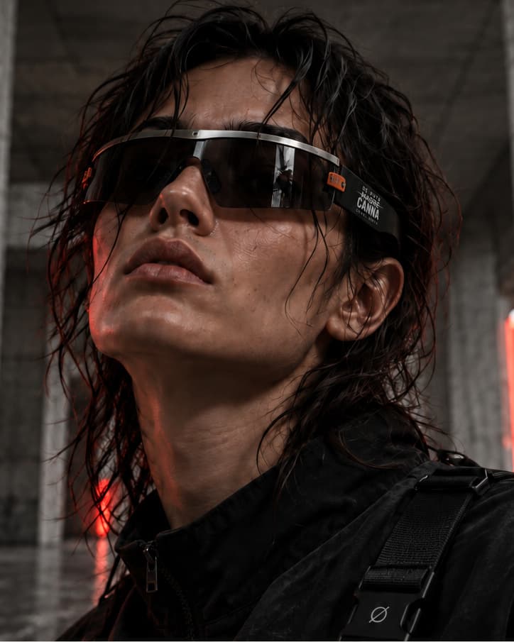 A model with wet hair in a smoked shield visor under a steel brow bar, looking up in a concrete hall lit red